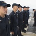 A group of U.S. Navy sailors in dark blue utility uniforms (NWU Type III or similar) are standing in formation on the deck of a naval vessel, likely during an inspection or muster. The ship appears to be underway at sea, visible from the ocean horizon in the background. One sailor on the far right appears to be holding a clipboard, suggesting an administrative or inspection activity.