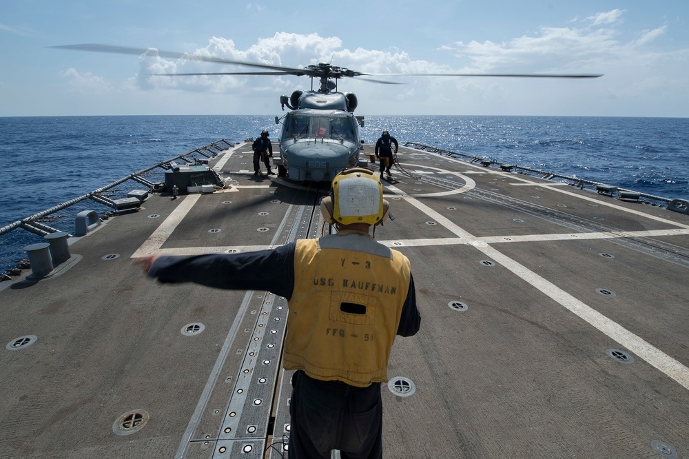 A naval flight deck crew member wearing a yellow vest marked 'USS KAUFFMAN FFG-51' signals on the helicopter landing deck of a U.S. Navy frigate. A military helicopter, likely an SH-60 Seahawk, sits on the flight deck with its rotors spinning while additional crew members attend to it. The scene takes place at sea under partly cloudy skies.