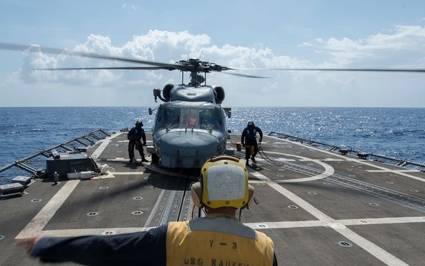 A naval flight deck crew member wearing a yellow vest marked 'USS KAUFFMAN FFG-51' signals on the helicopter landing deck of a U.S. Navy frigate. A military helicopter, likely an SH-60 Seahawk, sits on the flight deck with its rotors spinning while additional crew members attend to it. The scene takes place at sea under partly cloudy skies.