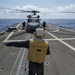 A naval flight deck crew member wearing a yellow vest marked 'USS KAUFFMAN FFG-51' signals on the helicopter landing deck of a U.S. Navy frigate. A military helicopter, likely an SH-60 Seahawk, sits on the flight deck with its rotors spinning while additional crew members attend to it. The scene takes place at sea under partly cloudy skies.