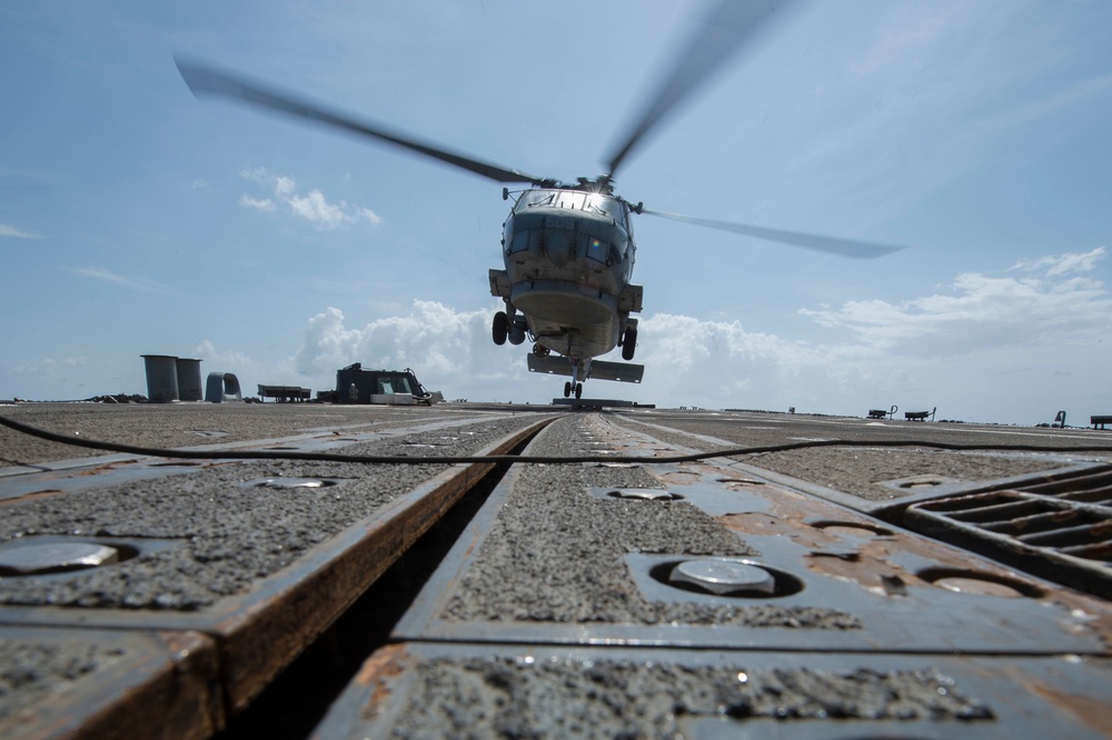 A military helicopter, appearing to be a Sikorsky MH-60 Seahawk variant, is captured in a low-angle shot as it hovers or lands on what appears to be a naval vessel's flight deck. The perspective is taken from ground level on the deck, emphasizing the textured metal surface with tie-down tracks and hardware in the foreground. The rotor blades are motion-blurred, indicating active rotation.