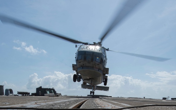 A military helicopter, appearing to be a Sikorsky MH-60 Seahawk variant, is captured in a low-angle shot as it hovers or lands on what appears to be a naval vessel's flight deck. The perspective is taken from ground level on the deck, emphasizing the textured metal surface with tie-down tracks and hardware in the foreground. The rotor blades are motion-blurred, indicating active rotation.