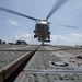 A military helicopter, appearing to be a Sikorsky MH-60 Seahawk variant, is captured in a low-angle shot as it hovers or lands on what appears to be a naval vessel's flight deck. The perspective is taken from ground level on the deck, emphasizing the textured metal surface with tie-down tracks and hardware in the foreground. The rotor blades are motion-blurred, indicating active rotation.