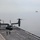 Osprey makes first-ever landing on Republic of Korea amphibious assault ship A V-22 Osprey tiltrotor aircraft sits on the flight deck of a naval vessel with its rotors spinning, indicating it is preparing for takeoff or has just landed. Several deck crew members in colored safety vests are visible on the flight deck. Multiple ships are visible in the background through hazy atmospheric conditions.