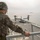 Osprey makes first ever landing on Republic of Korea amphibious assault ship A soldier in digital camouflage uniform stands on an elevated observation platform of a naval vessel, observing a tiltrotor aircraft (V-22 Osprey) on the ship's flight deck below. The scene appears to be at sea, with a second vessel visible on the horizon in hazy conditions. This appears to be a military naval operation or exercise.