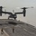 Osprey makes first ever landing on Republic of Korea amphibious assault ship A V-22 Osprey tiltrotor aircraft is landing or taking off from the flight deck of a naval vessel, likely an amphibious assault ship. Two flight deck crew members in yellow and gray jerseys are visible on the deck, with one directing the aircraft. A second naval vessel is visible in the background on the open water.