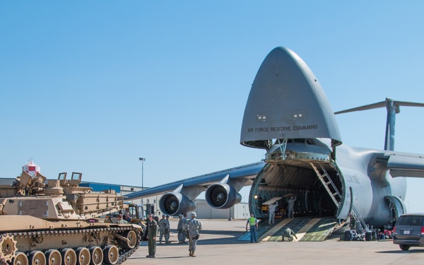 A military loading operation is underway on an airfield tarmac, where an M1 Abrams main battle tank in desert tan camouflage is positioned near a large C-5 Galaxy military transport aircraft with its nose cargo door raised. The aircraft bears 'Air Force Reserve Command' markings on its fuselage. Multiple military personnel are visible around both vehicles, suggesting an active loading or unloading exercise or deployment operation.