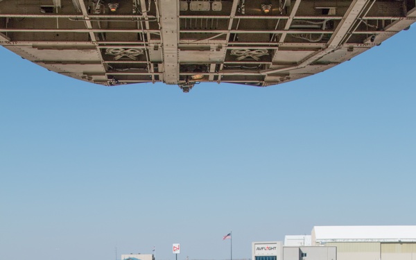 A military loadmaster in a flight suit stands on the rear cargo ramp of a large military transport aircraft, using hand signals to guide M1 Abrams main battle tanks onto the aircraft at a civilian/military airfield. The perspective is from inside the aircraft looking outward, with the open cargo ramp and overhead fuselage structure framing the scene. A second military personnel member is visible walking on the tarmac in the background near an 'AVFLIGHT' FBO facility.