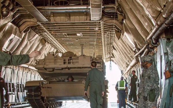 Military personnel are loading or unloading a tracked armored vehicle, likely an M2 Bradley Infantry Fighting Vehicle, into or out of a large military cargo aircraft, consistent with a C-17 Globemaster III. Several individuals in flight suits and Army combat uniforms are guiding the vehicle along the cargo bay floor. A safety marshal in a high-visibility vest is visible near the rear ramp opening.