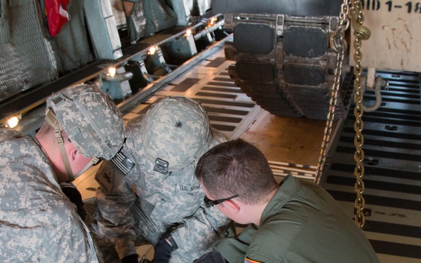 Three military personnel are crouched on the cargo floor of a large military transport aircraft, appearing to work on securing equipment or rigging. Two individuals are wearing U.S. Army ACU (Army Combat Uniform) camouflage pattern, while the third wears an olive green flight suit with a U.S. flag patch. They appear to be collaborating on a task involving tie-down chains or cargo restraint hardware.