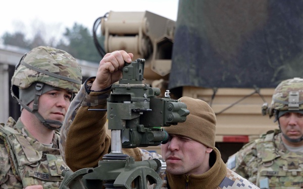 Three military personnel are gathered around what appears to be a crew-served weapon or artillery piece during what looks like a training or demonstration exercise. Two soldiers in US Army MultiCam/OCP uniforms flank a third individual wearing digital camouflage and a brown beanie, who is looking through an optical sight. A military tactical vehicle is visible in the background.