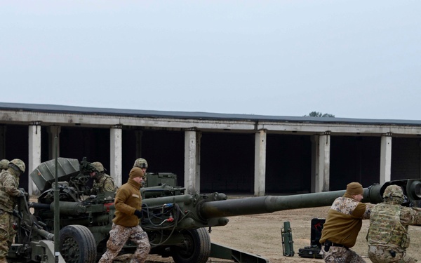 Military personnel in mixed uniforms are operating a large towed artillery piece, likely an M777 or M198 howitzer, on a sandy training ground. Soldiers wearing both standard multicam/OCP uniforms and Flecktarn (German camouflage) pattern uniforms are working together, suggesting a multinational training exercise. The activity appears to be emplacement or repositioning of the artillery system near a large industrial or military building.