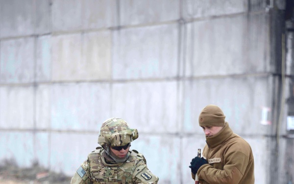 Two military personnel are working together in an outdoor area near a concrete structure, appearing to conduct a ground survey or lane-marking exercise. One soldier is in full US Army MultiCam/OCP uniform with helmet and body armor, while the second individual wears a tan fleece jacket with digital camouflage trousers, suggesting a multinational training exercise. They are handling measuring poles with orange-and-black marker stakes connected by white tape or cord.