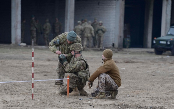 Military personnel are engaged in what appears to be a training exercise on a sandy, open ground near industrial or warehouse-style buildings. Several soldiers in multicam/camouflage uniforms are clustered around a marked area with orange and red-white striped poles and a rope/tape boundary, suggesting mine clearance or IED detection training. A blurred soldier in the foreground is in a prone or low position, also participating in the exercise.