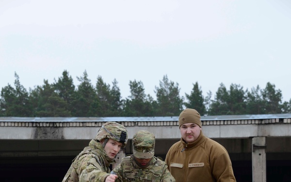 Three military personnel are gathered on a sandy training ground, with two in U.S. Army multicam uniforms and one in a tan fleece jacket with digital camouflage pants bearing a name tag reading 'A. STREKNUVS'. They appear to be conducting a joint training exercise, focused on an object or tool being handled near the ground. An orange and black marker pole is visible in the foreground, and a concrete structure with trees is visible in the background.