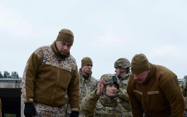 A group of military personnel in mixed uniforms are gathered outdoors on a training ground. A soldier identified by name tape as 'MCALLISTER' in multicam/OCP uniform and helmet is kneeling and gesturing while holding a marking stake with orange and white tape, appearing to brief or instruct others. Surrounding personnel are wearing what appears to be Latvian digital camouflage uniforms, suggesting a multinational military training exercise.