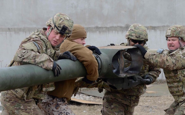 Four military personnel in mixed camouflage uniforms are collaboratively lifting or maneuvering a large green cylindrical object, likely a tube or barrel component of heavy equipment. At least two distinct camouflage patterns are visible — one consistent with US Army Operational Camouflage Pattern (OCP) and one consistent with German Bundeswehr Flecktarn pattern — suggesting a multinational military exercise or operation. The setting appears to be an outdoor military compound or training area.