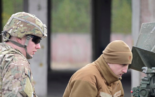 Two military personnel are working on what appears to be an artillery piece or towed weapon system. One soldier is in full combat gear including a helmet and body armor, while the other wears a tan/coyote brown fleece and beanie cap. The second individual appears to be operating or adjusting the weapon's sighting and breech mechanisms.