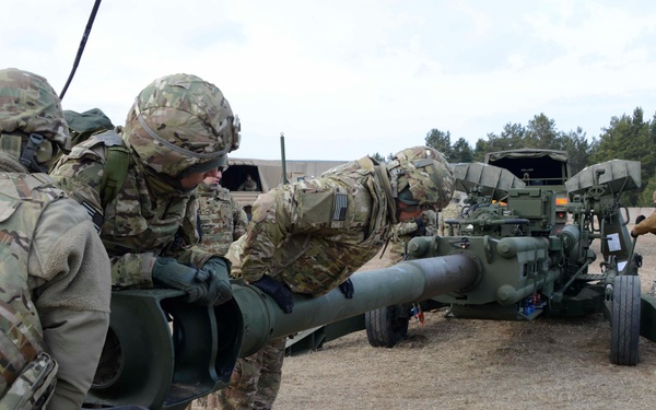 Military personnel in camouflage uniforms are working together to position or maneuver an artillery piece, likely an M777 howitzer, on a sandy training ground. Multiple soldiers in US Army MultiCam/OCP uniforms are physically handling the barrel and trail of the howitzer, while additional personnel in different uniforms observe nearby. The setting appears to be a military training exercise at an outdoor range surrounded by conifer trees.