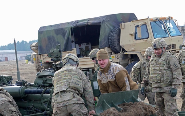 A group of military personnel in camouflage uniforms are working together to set up or position what appears to be an M777 or similar towed artillery piece on sandy/muddy terrain. Several soldiers are physically handling a spade or trail component of the artillery system, with one individual in a different uniform (possibly a foreign military counterpart) assisting. A military tactical truck (likely a FMTV/LMTV) is visible in the background, suggesting a field exercise or deployment operation.
