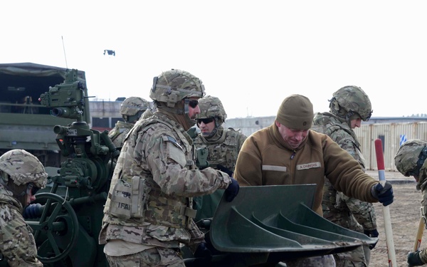 A group of military personnel in camouflage uniforms are working together to set up or position what appears to be an artillery piece, likely an M777 howitzer or similar field gun. The soldiers are working in a forward operating area with military vehicles visible in the background. One individual wearing a tan fleece jacket and digital camouflage trousers, possibly a foreign military advisor or liaison, is assisting the predominantly US Army soldiers.