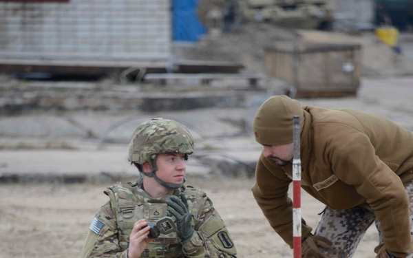 Two military personnel are working together in a sandy outdoor area, examining a marked point on the ground near a red-and-white measuring/survey pole and an orange stake with white tape. One soldier is in full US Army multicam uniform with helmet and body armor, kneeling and holding a device. The second individual wears a digital camouflage uniform with a tan fleece top and beanie, leaning over to inspect the marked area.