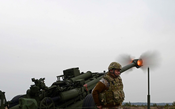 Military personnel are operating a large artillery piece, captured at the moment of firing with a visible muzzle blast and flame at the barrel. At least two distinct uniform patterns are visible — US Army MultiCam/OCP and what appears to be German Flecktarn camouflage — suggesting a multinational training exercise. The setting is an open, flat, sparsely vegetated field under an overcast sky.