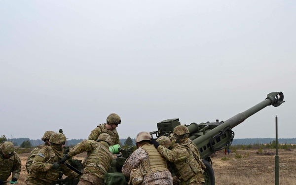 A group of military personnel in mixed camouflage uniforms are working together to operate or position a large artillery piece, likely an M777 howitzer or similar towed howitzer, in an open field training environment. The soldiers appear to represent at least two different nations based on their distinct camouflage patterns. The setting appears to be a military training exercise on flat, sparse terrain with a treeline visible in the background.