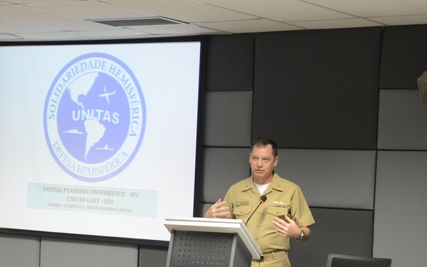 A military officer in U.S. Navy tan/khaki service uniform is presenting at a podium during the UNITAS LANT-LVI Initial Planning Conference (IPC). The presentation screen displays the UNITAS 'Solidariedade Hemisférica / Defesa Hemisférica' logo along with conference details. An audience of military personnel from multiple nations is seated in a conference room setting.