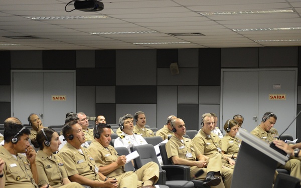 A military briefing or lecture is taking place in a conference room where a civilian or official in dark clothing is presenting at a podium to a large audience of uniformed military personnel. The audience members are wearing tan/khaki uniforms and many are equipped with translation headsets, suggesting a multilingual or international meeting. Exit signs reading 'SAIDA' (Portuguese for 'exit') indicate this event is taking place in a Portuguese-speaking country, likely Brazil.