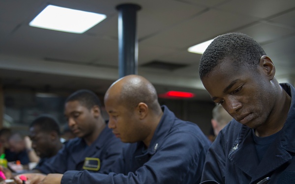 A group of individuals wearing matching dark navy blue uniforms are seated at a long wooden table, focused on writing or marking documents on clipboards. The setting appears to be an indoor facility, possibly a military mess hall or common area. Multiple people are engaged in what appears to be a standardized task, such as filling out forms or taking a test.