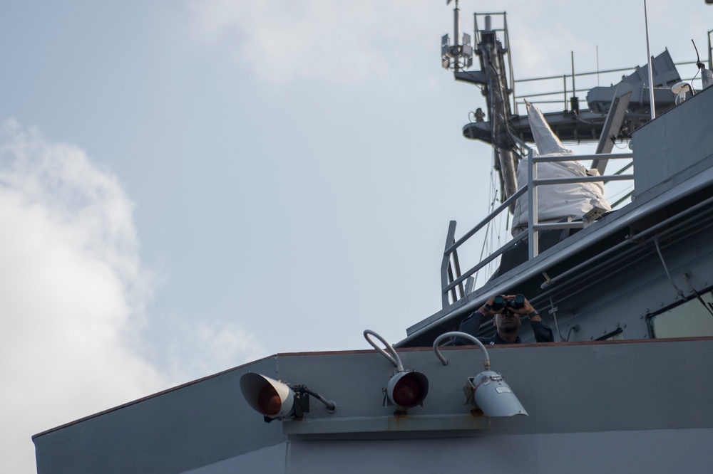 A naval vessel is shown from a low-angle perspective, with a crew member using binoculars from an elevated deck position. The ship's superstructure, mast with communications equipment, railings, and deck-mounted lights and fittings are clearly visible. The image captures a watchkeeping or observation activity aboard what appears to be a military or coast guard ship.