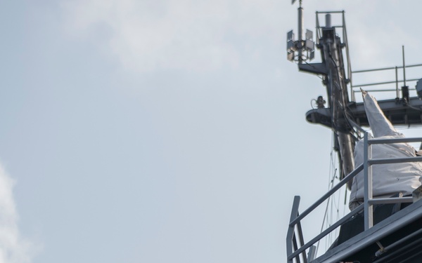 A naval vessel is shown from a low-angle perspective, with a crew member using binoculars from an elevated deck position. The ship's superstructure, mast with communications equipment, railings, and deck-mounted lights and fittings are clearly visible. The image captures a watchkeeping or observation activity aboard what appears to be a military or coast guard ship.