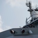 A naval vessel is shown from a low-angle perspective, with a crew member using binoculars from an elevated deck position. The ship's superstructure, mast with communications equipment, railings, and deck-mounted lights and fittings are clearly visible. The image captures a watchkeeping or observation activity aboard what appears to be a military or coast guard ship.