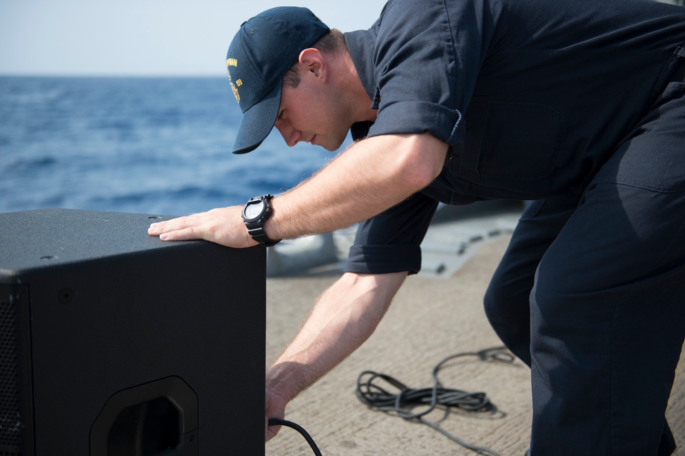 A naval service member dressed in dark navy working uniform and a ship-name cap is leaning over a large black speaker cabinet on the deck of a warship. He appears to be connecting or adjusting a cable at the rear of the speaker. The ocean is visible in the background, confirming an at-sea setting.