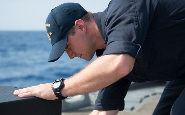 A naval service member dressed in dark navy working uniform and a ship-name cap is leaning over a large black speaker cabinet on the deck of a warship. He appears to be connecting or adjusting a cable at the rear of the speaker. The ocean is visible in the background, confirming an at-sea setting.