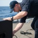 A naval service member dressed in dark navy working uniform and a ship-name cap is leaning over a large black speaker cabinet on the deck of a warship. He appears to be connecting or adjusting a cable at the rear of the speaker. The ocean is visible in the background, confirming an at-sea setting.