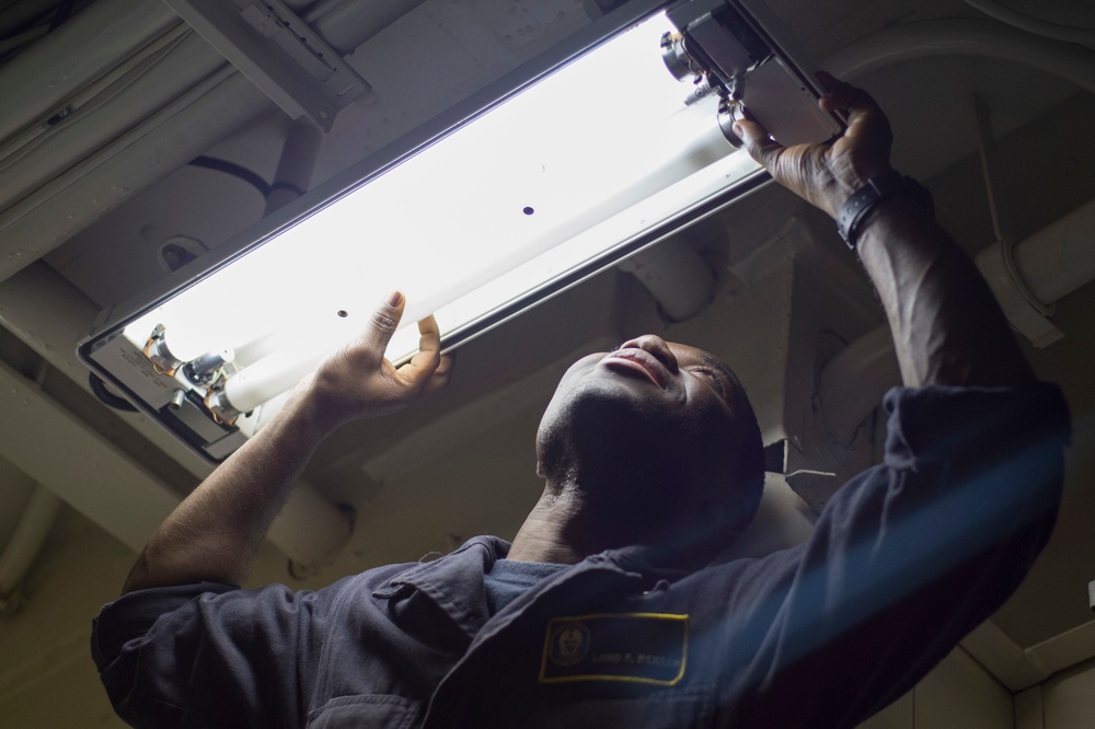 A person in a dark blue uniform with a name tag and insignia patch is working overhead, replacing or installing a fluorescent light fixture. The photo is taken from a low angle looking upward, showing the individual using both hands to manipulate the light tube and its mounting hardware. The setting appears to be an interior space with a metal ceiling, possibly a military vessel or institutional building.