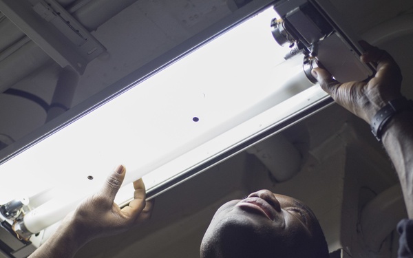 A person in a dark blue uniform with a name tag and insignia patch is working overhead, replacing or installing a fluorescent light fixture. The photo is taken from a low angle looking upward, showing the individual using both hands to manipulate the light tube and its mounting hardware. The setting appears to be an interior space with a metal ceiling, possibly a military vessel or institutional building.