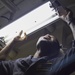 A person in a dark blue uniform with a name tag and insignia patch is working overhead, replacing or installing a fluorescent light fixture. The photo is taken from a low angle looking upward, showing the individual using both hands to manipulate the light tube and its mounting hardware. The setting appears to be an interior space with a metal ceiling, possibly a military vessel or institutional building.