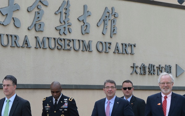 A group of six men are walking in front of the Okura Museum of Art in Tokyo, Japan. The group includes one individual in a U.S. Army dress uniform and five men in dark business suits. The setting appears to be an official or diplomatic visit based on the formal attire and coordinated movement.