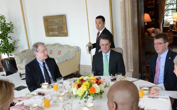 A formal dining meeting is taking place around a large round table set with fine china, gold-rimmed plates, and a floral centerpiece. The group includes civilians in business attire and at least one military officer in dress uniform. The setting appears to be an upscale official residence or government dining room.