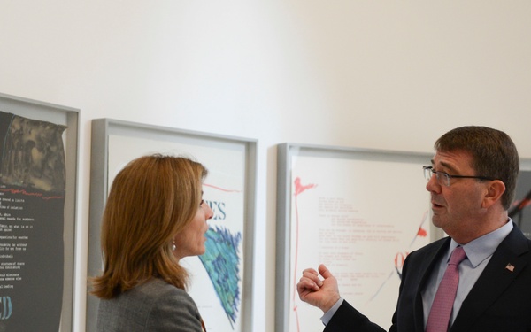 Two individuals are engaged in conversation in what appears to be an art gallery or exhibition space. A man in a dark suit with a pink tie and glasses is speaking to a woman in a gray blazer, both standing in front of framed artwork on a white wall. The artworks appear to feature text and abstract imagery, with one piece visibly containing the word 'UNITED'.