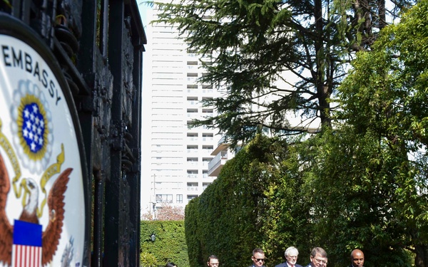 A group of approximately six individuals, including civilians in business attire and at least one military officer in uniform, are walking along a pathway at the Embassy of the United States of America. The U.S. Embassy seal is prominently displayed on a black iron gate in the foreground. The setting appears to be a well-maintained embassy compound with trees and a modern building visible in the background.