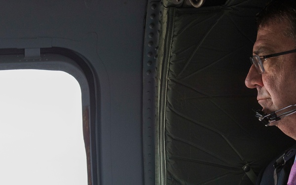 A man in business attire is seated inside a military-style helicopter, looking out the window at an urban landscape below. He is wearing olive-green aviation headphones with a microphone and holding printed documents. The aerial view suggests the aircraft is flying at significant altitude over a densely populated city.