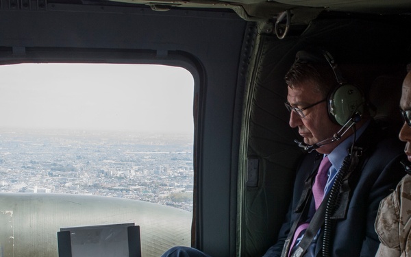 Three individuals are seated inside a military helicopter in flight, with an aerial view of a coastal city visible through the window. A civilian in a dark suit and pink tie is reviewing a document, flanked by two individuals wearing military-style flight headsets. The scene suggests an official aerial reconnaissance or transport mission.