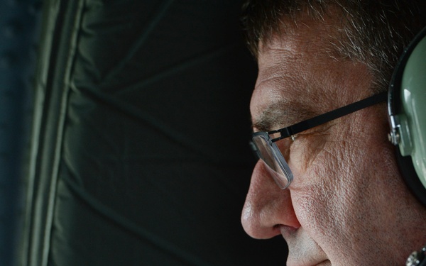 A close-up profile shot of a middle-aged man wearing rectangular-framed glasses and a military-style olive green aviation headset with a boom microphone. He appears to be inside an aircraft, looking outward through what appears to be a window or opening. The lighting is dramatic, illuminating his face from the front while the background remains dark.