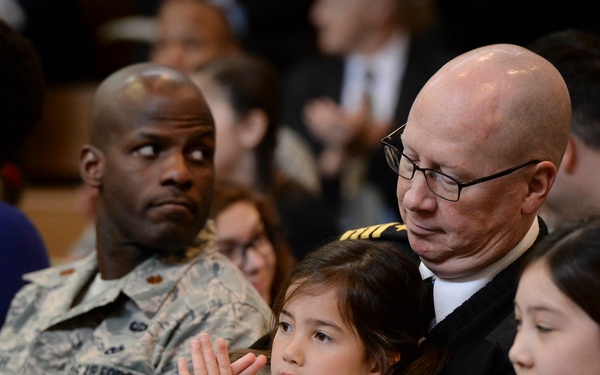 A formal military or official ceremony is taking place with uniformed personnel and civilians seated in an audience. A bald man in a dark naval uniform holds a young girl on his lap, while others around them are applauding. The scene suggests a solemn or ceremonial event attended by military members and their families.