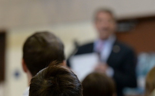 A group of people, including children and adults, are seated in rows facing a speaker at the front of a room. The speaker, a man in a dark suit, is holding papers and appears to be addressing the audience. The scene suggests a formal or semi-formal presentation, ceremony, or meeting.