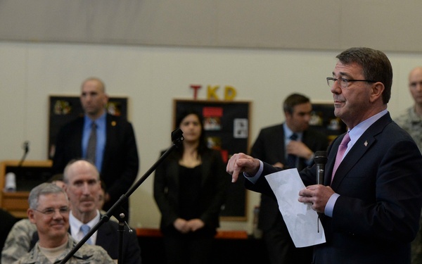 A man in a dark suit is delivering a speech at a microphone stand inside what appears to be a military gymnasium or recreational facility, holding papers and a microphone. The audience includes military personnel in camouflage uniforms, civilians, and security personnel standing in the background. The setting suggests an official military briefing or town hall event.